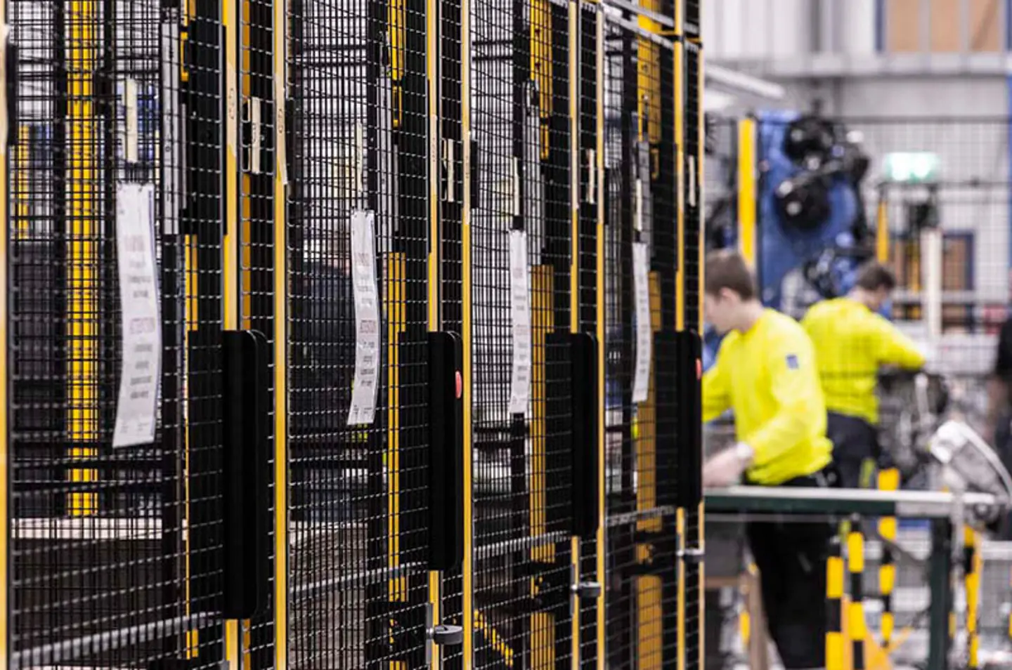Travailleurs dans une usine, à côté de barrières de sécurité noires et jaunes avec panneaux grillagés métalliques.